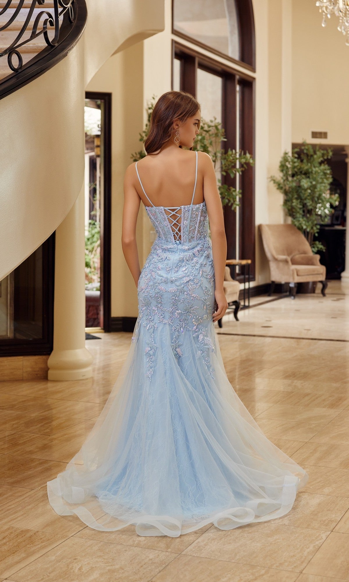 A woman in the Nox Anabel H1874, a light blue long prom dress with trumpet silhouette, lace details, and thin straps, stands indoors on a polished marble floor near a staircase, facing away.